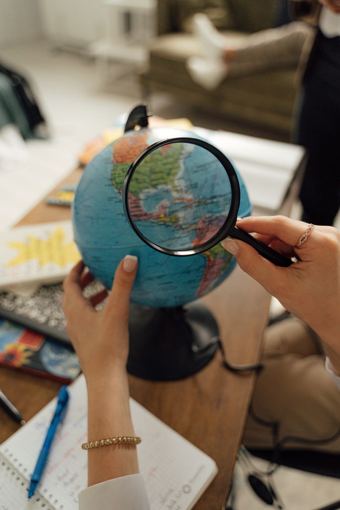 Close-up of hands using a magnifying glass to examine a globe on a desk with notes around.