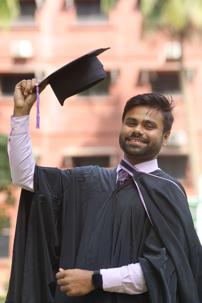 Smiling graduate wearing a gown and holding a mortarboard outdoors on graduation day.